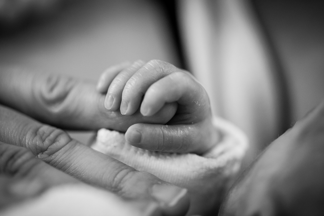 Black and white picture of the hand of a tiny baby wrapped around the finger of a parent.