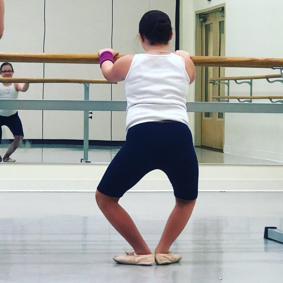 Back of girl with Down syndrome doing ballet, holding to bar and doing a plie, you can see her reflection in the mirror. She is wearing a white shirt, black shorts and ballet slippers.
